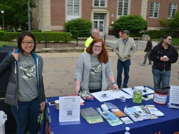 A plaza outdoors where two smiling people facing camera stand behind a table covered with brochures, etc., with other people standing around behind