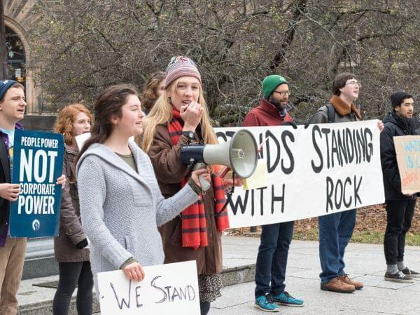 Seven college students, dressed for cold, stand in a line at a rally. One speaks into a megaphone and others hold signs.