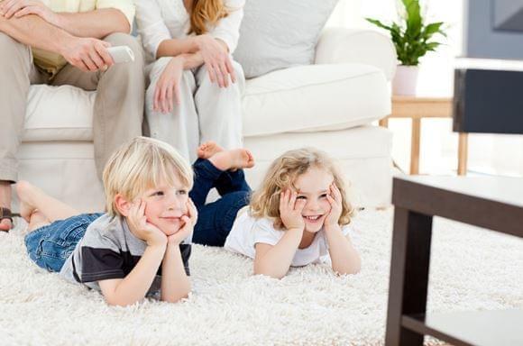 2 small children laying on floor watching tv. Parents are seated on couch.