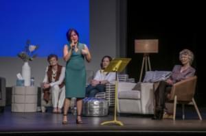 Woman stands on stage with microphone with three women sitting behind her