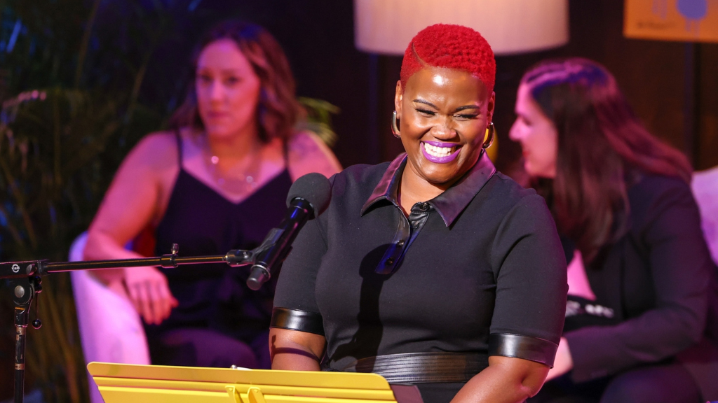 Woman stands on stage with microphone, other women sit behind her