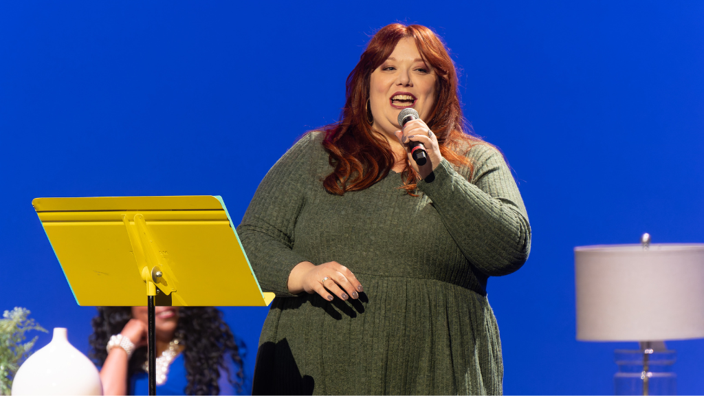 Woman stands on stage with microphone, other women sit behind her