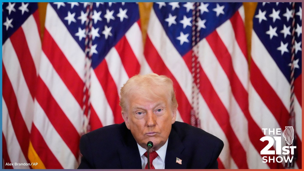 Donald Trump sitting before a microphone, with a wall of American flags behind him