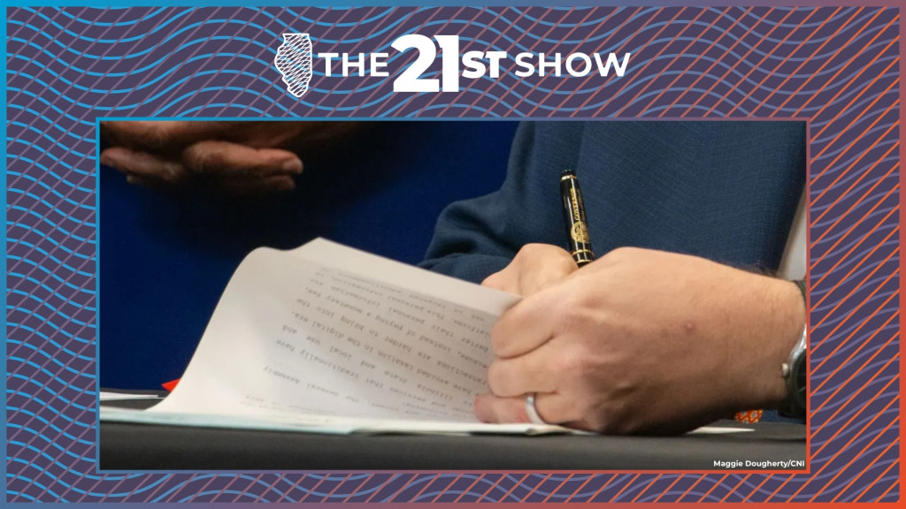 a close up view of a piece of paper, held by Pritzker's left hand (which features a silver-colored wedding band) while he uses his right hand to sign the papers; the pen is black with gold accents, including the seal of the state of Illinois and the word GOVERNOR