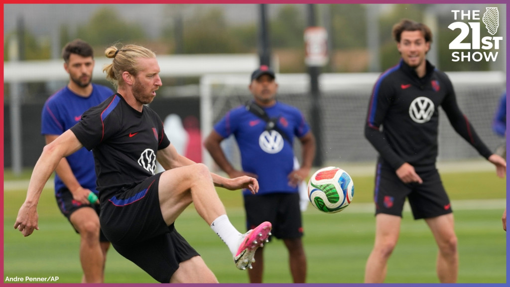 a white man with blonde hair pulled back into a man-bun kicks the red, green, blue and white soccer ball fo the World Cup. He's wearing a black outfit with a the US soccer logo, the Nike swoosh, and the VW logo on 