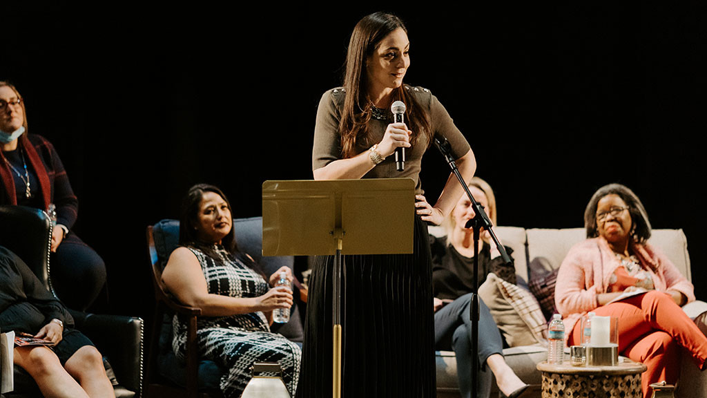 Woman stands on stage with a microphone with people behind her on couches