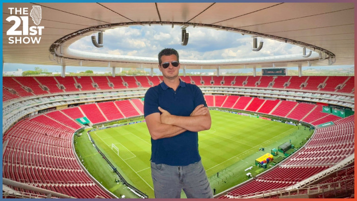 A white man wearing aviator shades, a navy blue polo shirt, and jeans stands with his arms crossed near the top of Guadalajara Stadium, the bowl-shaped inside of which can be seen behind him. The seats are red, the pitch is green, and there's a white awning over all the seating area.