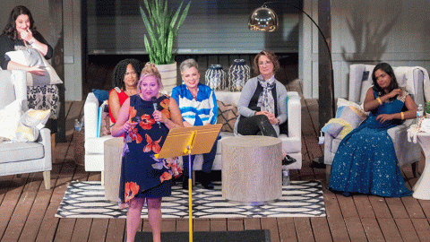 woman on stage talking with other women sitting on couches and chairs behind her