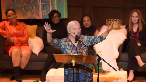 Woman on stage behind a music stand with hands raised speaking to a crowd