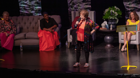 Woman standing on stage holding a microphone talking to a crowd