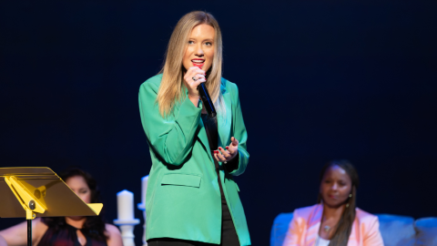 Woman on stage in green blazer holding microphone. Two woman sit behind her on couch.