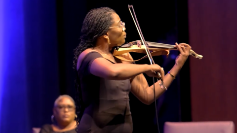 Woman on stage holding and playing violin or viola. Woman behind her. 