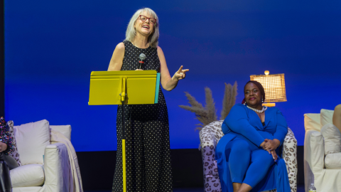 Woman stands on stage with microphone, other women sit behind her