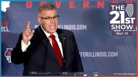 A man with close-cropped, graying hair wearing a dark blue suit with a white dress shirt and red tie. He raises the index finger on his right hand as he speaks. Behind him is a backdrop with his name, web address, and logo of his campaign.