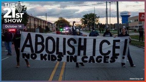 at sunset, three women in dark clothing hold a white banner with huge black lettering. It reads, 