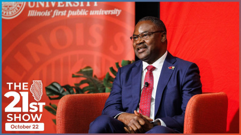 a Black man wearing a blue suit, white shirt and red necktie sits in an orange lounge chair. Behind him is an indoor plant and large red banners with Illinois State University's logo