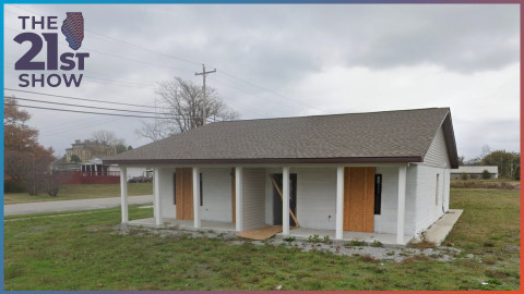 A small white house with plywood over the doors and windows is in a patchy field of grass under overcast skies.
