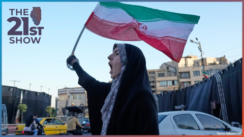 a woman wears a lavander, floral-print hijab under a black chador as she waves a green, white and red flag; she is standing along what appears to be a busy road, and her mouth is open wide, suggesting she is shouting