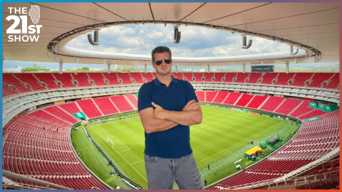 A white man wearing aviator shades, a navy blue polo shirt, and jeans stands with his arms crossed near the top of Guadalajara Stadium, the bowl-shaped inside of which can be seen behind him. The seats are red, the pitch is green, and there's a white awning over all the seating area.