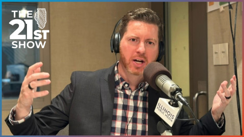 a white man in his 40s with brown hair and a beard is wearing a brown sport coat over a plaid button-down shirt; he's wearing headphones and taking into a microphone in a radio studio