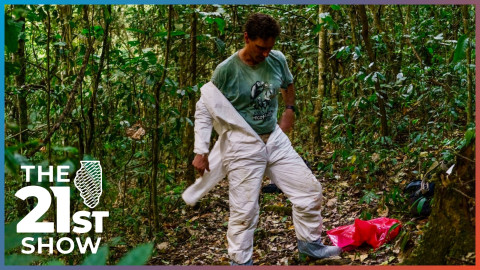 a white man in green T-shirt pulls on a white biohazard suit; there are light blue booties over his shoes and a red biohazard bag lies next to him on a bed of leaves in what appears to be a dense jungle
