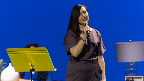 Woman stands on stage with microphone, other women sit behind her