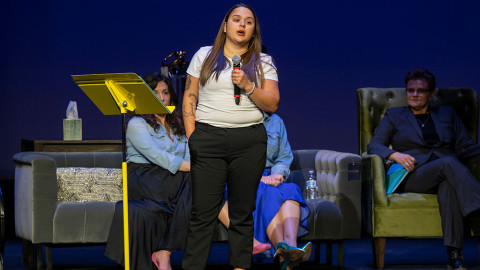 Woman stands on stage with microphone, others sit on couches behind her